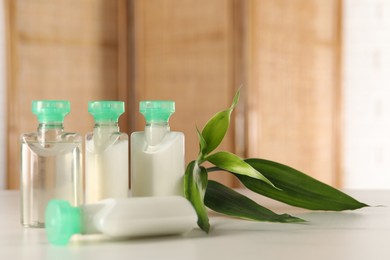 Photo of Mini bottles of cosmetic products and green branch on white table against blurred background. Space for text