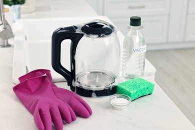 Photo of Cleaning electric kettle. Bottle of vinegar, sponge, rubber gloves and baking soda on countertop in kitchen