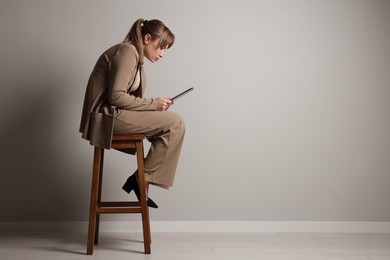 Photo of Young woman with poor posture using tablet while sitting on stool near grey wall, space for text