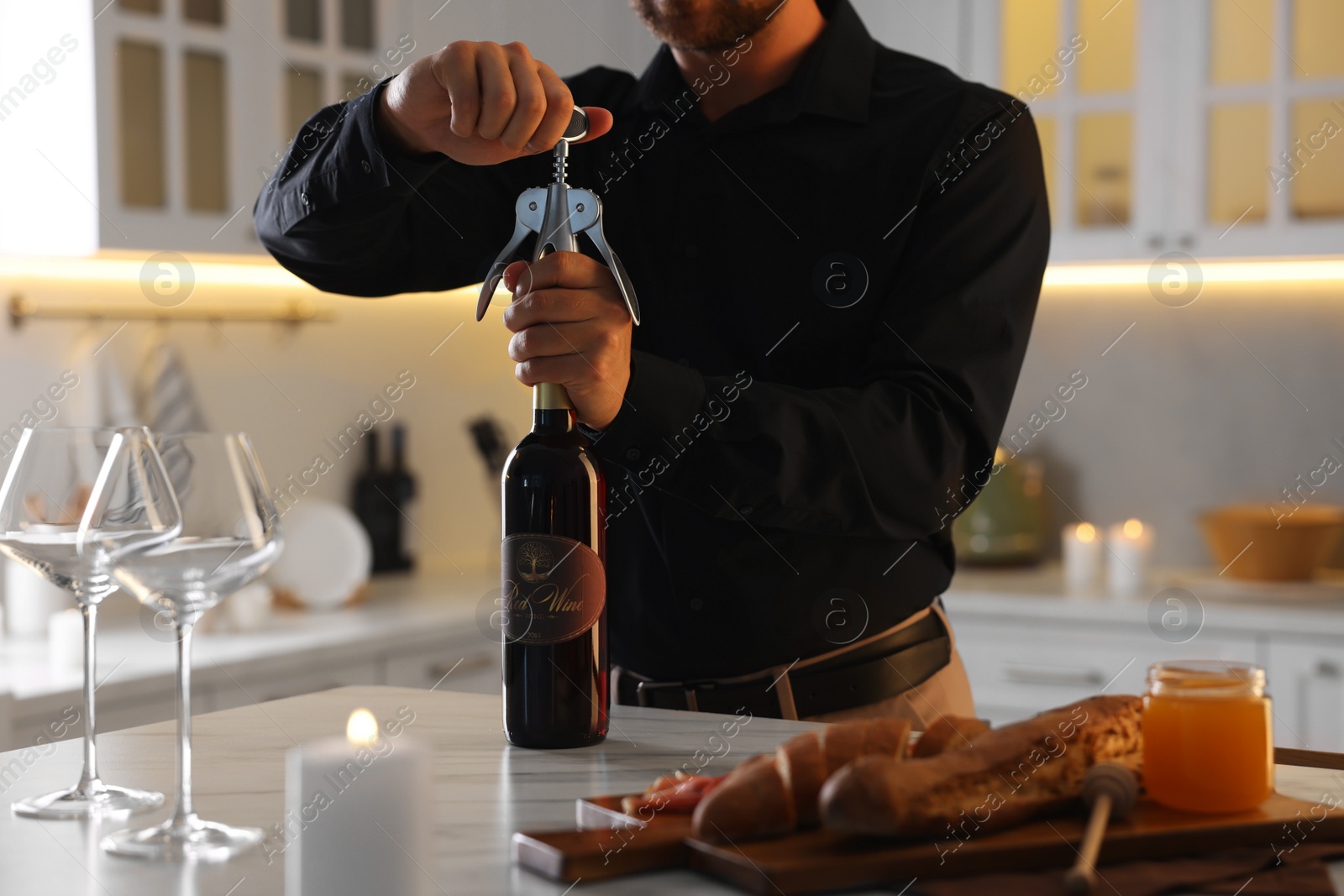 Photo of Romantic dinner. Man opening wine bottle with corkscrew at table in kitchen, closeup