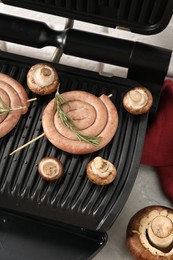 Photo of Electric grill with homemade sausages, rosemary and mushrooms on marble table, above view