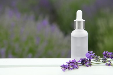 Bottle of essential oil and lavender flowers on white wooden table in field, space for text