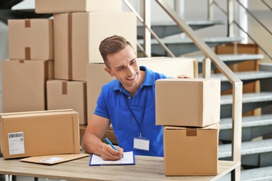 Photo of Young courier working with papers among parcels at table in delivery department