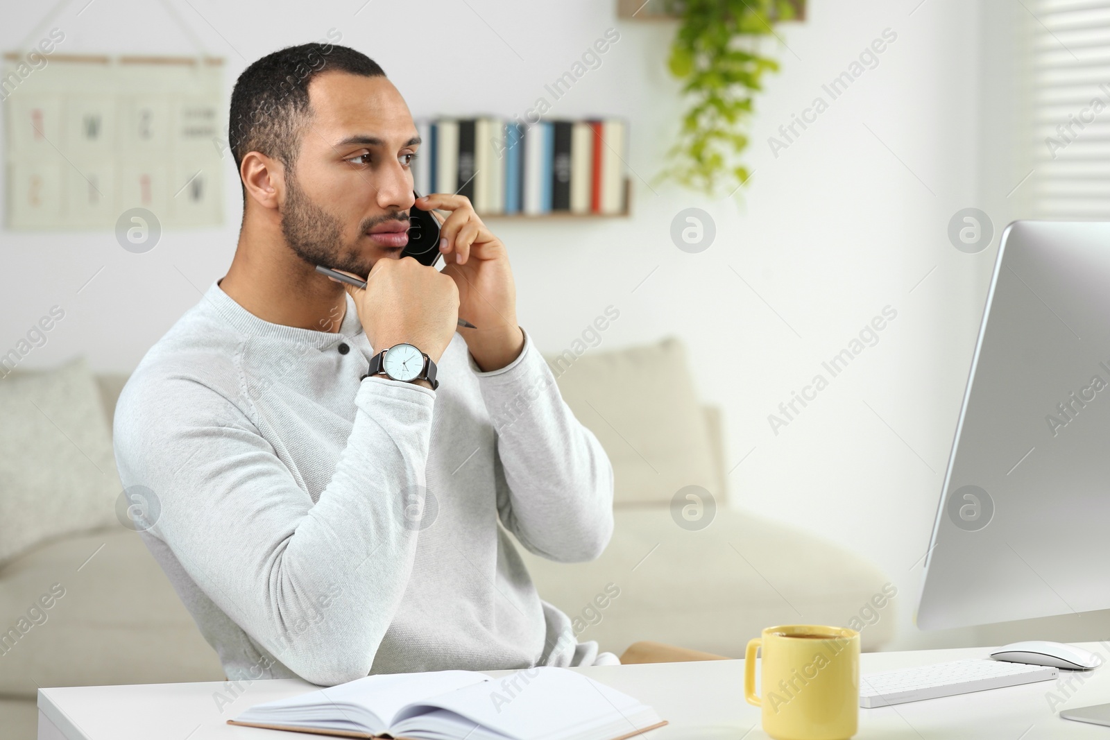 Photo of Young man talking on smartphone while working with computer at desk in room. Home office