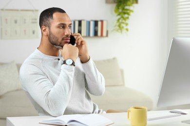 Young man talking on smartphone while working with computer at desk in room. Home office