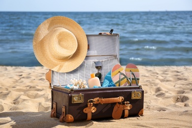 Open suitcase with stylish beach accessories on sand near sea