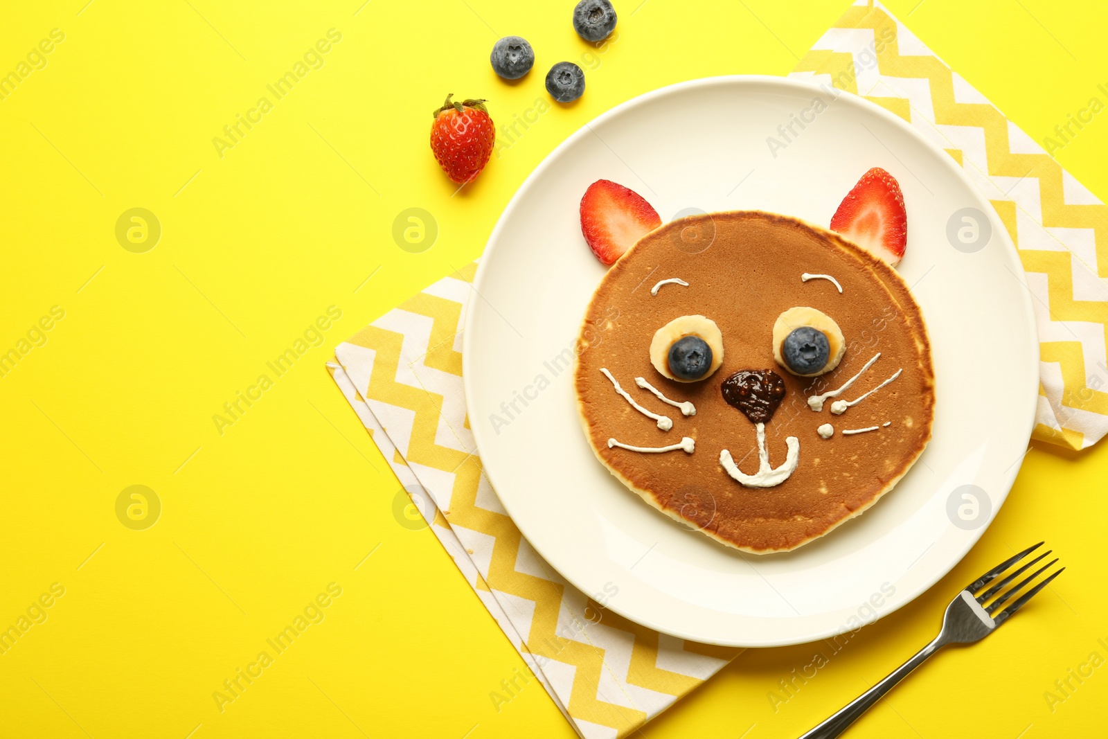 Photo of Creative serving for kids. Plate with cute cat made of pancakes, berries, cream, banana and chocolate paste on yellow background, flat lay. Space for text