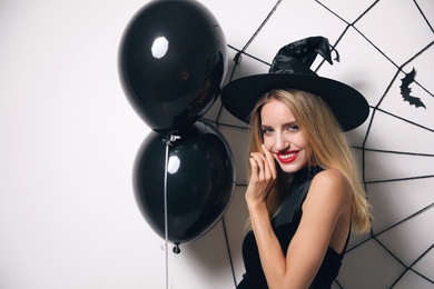 Woman in witch hat with balloons posing near white wall decorated for Halloween