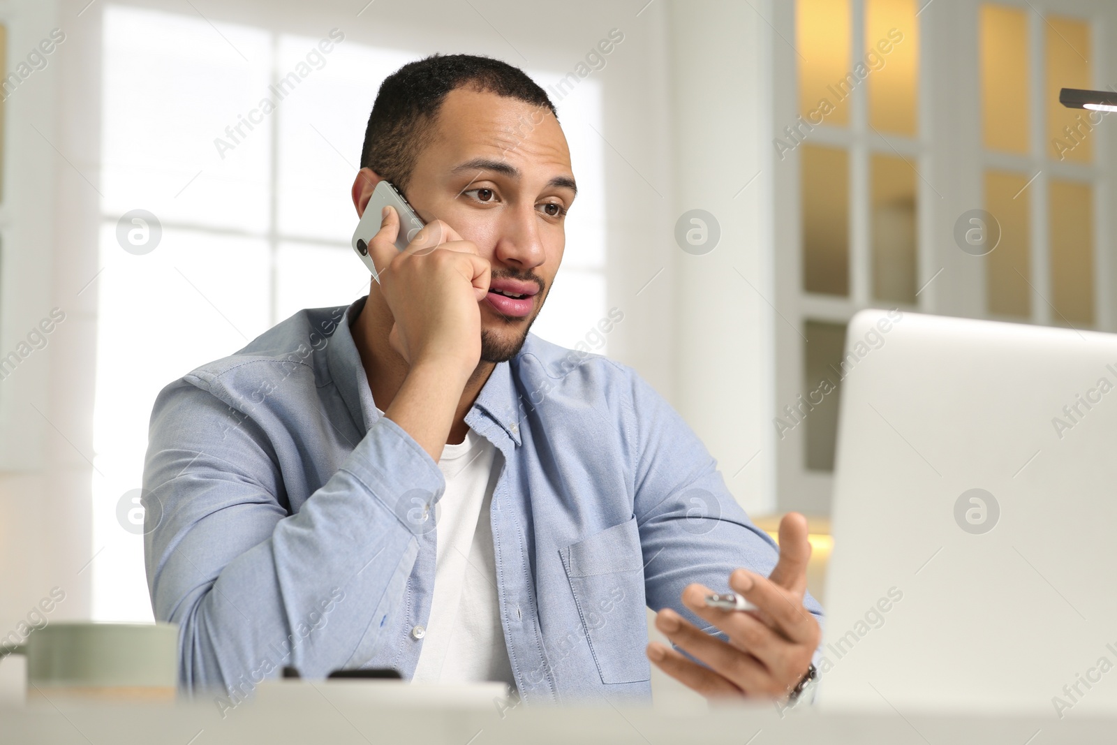 Photo of Young man talking on smartphone while working with laptop at desk in kitchen. Home office