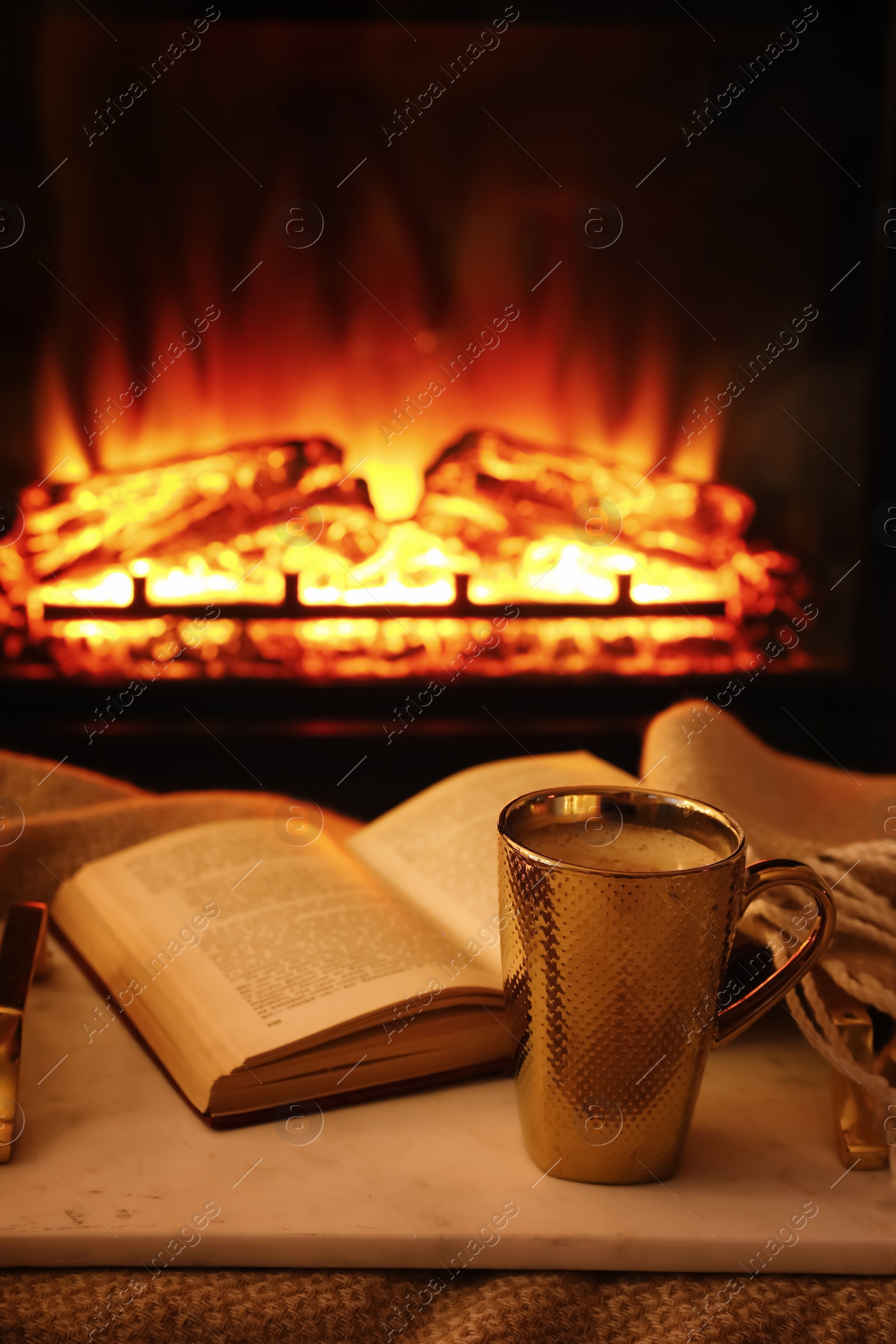 Photo of Cup of hot drink and book on blanket near fireplace indoors. Magic atmosphere