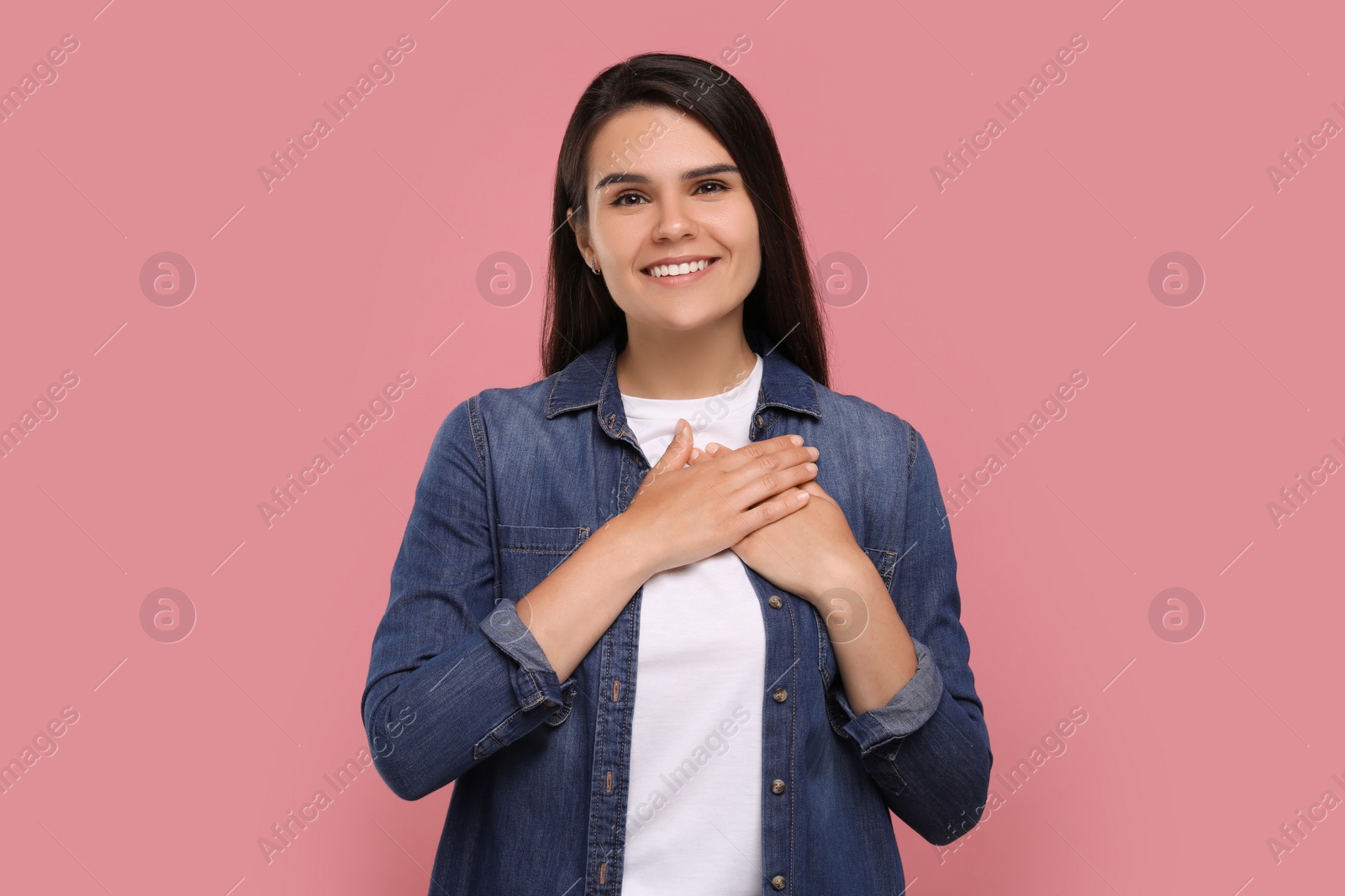 Photo of Thank you gesture. Beautiful grateful woman holding hands near heart on pink background