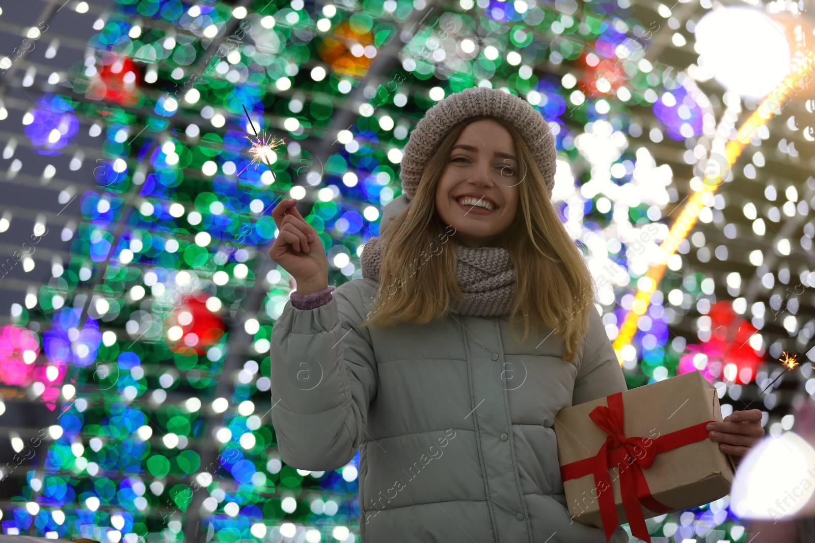 Photo of Happy young woman with gift box near festive lights. Christmas celebration