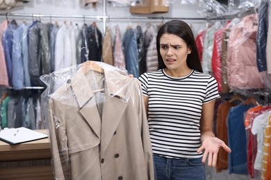 Photo of Dry-cleaning service. Displeased woman holding hanger with coat in plastic bag indoors