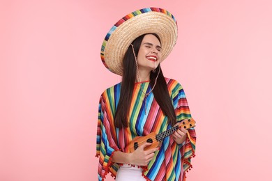 Young woman in Mexican sombrero hat and poncho playing ukulele on pink background