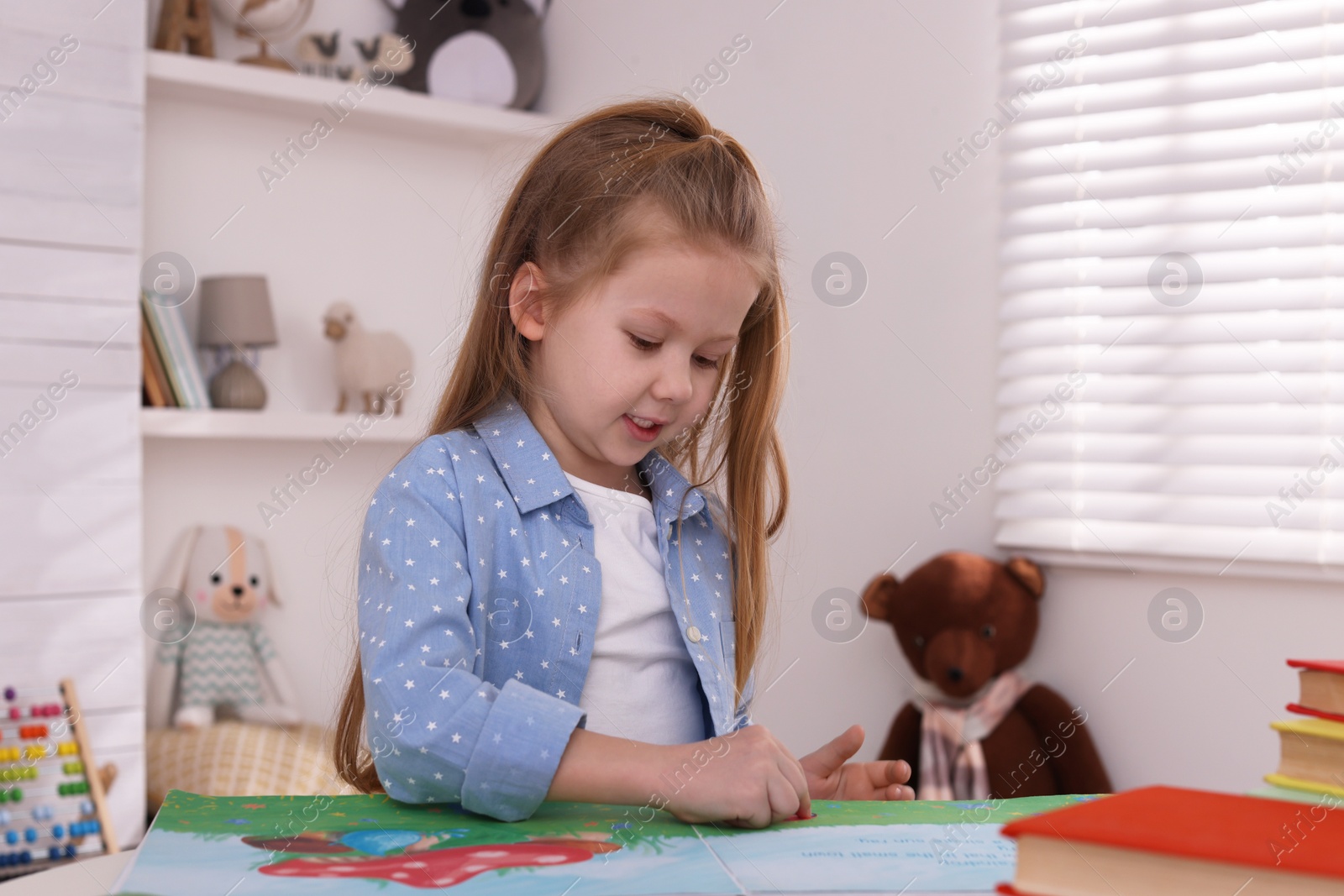 Photo of Cute little girl reading book at table in room