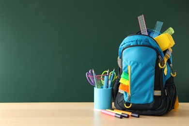 Photo of Stylish backpack and different school stationery on wooden table near green chalkboard, space for text. Back to school