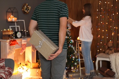 Man hiding gift box while his girlfriend decorating Christmas tree at home