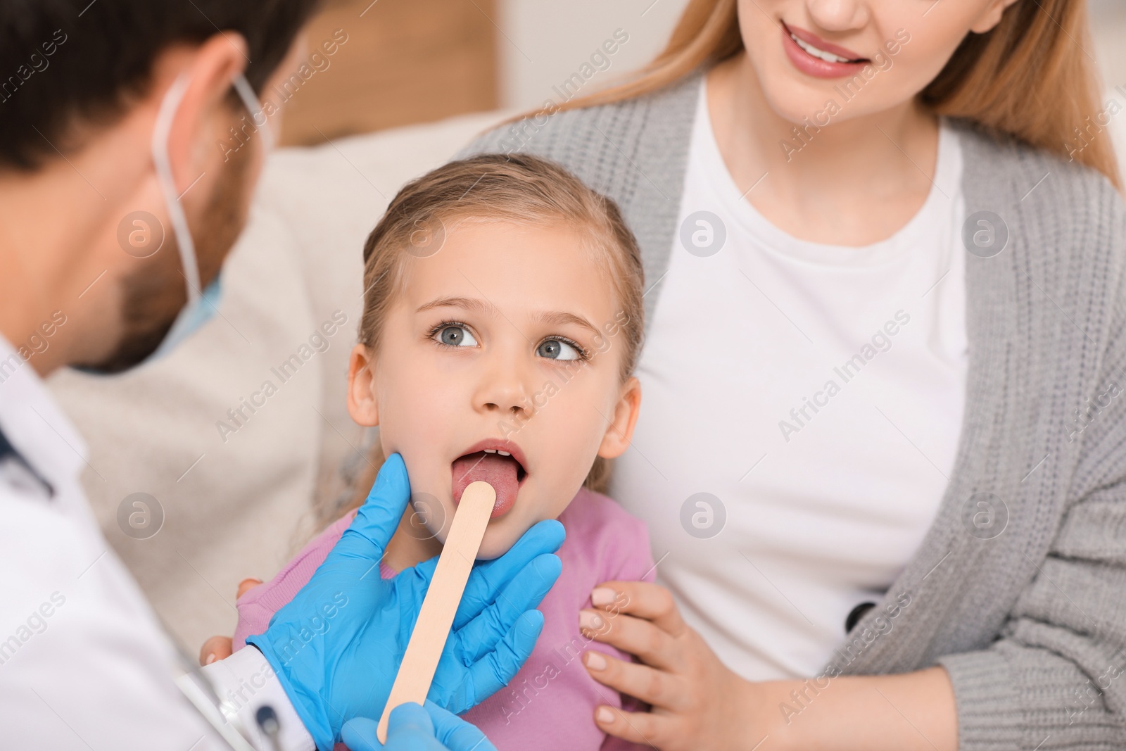 Photo of Doctor in medical mask examining girl`s oral cavity with tongue depressor near her mother indoors, closeup