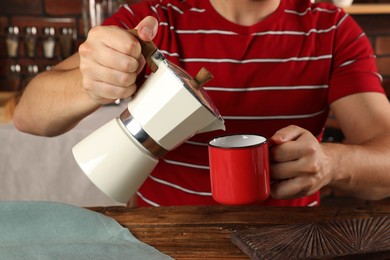 Man pouring aromatic coffee from moka pot into mug at wooden table indoors, closeup