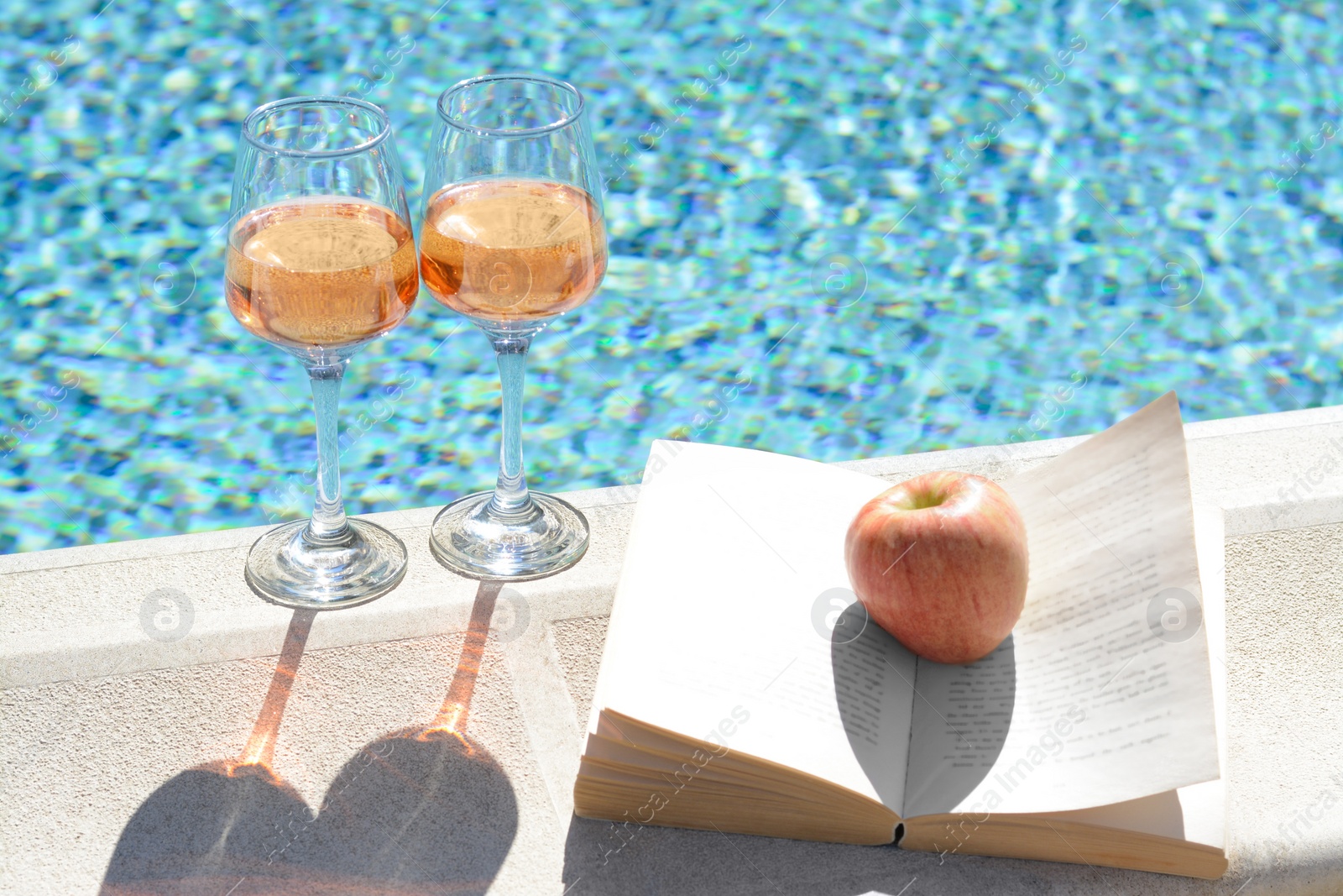 Photo of Glasses of tasty rose wine, open book and apple on swimming pool edge