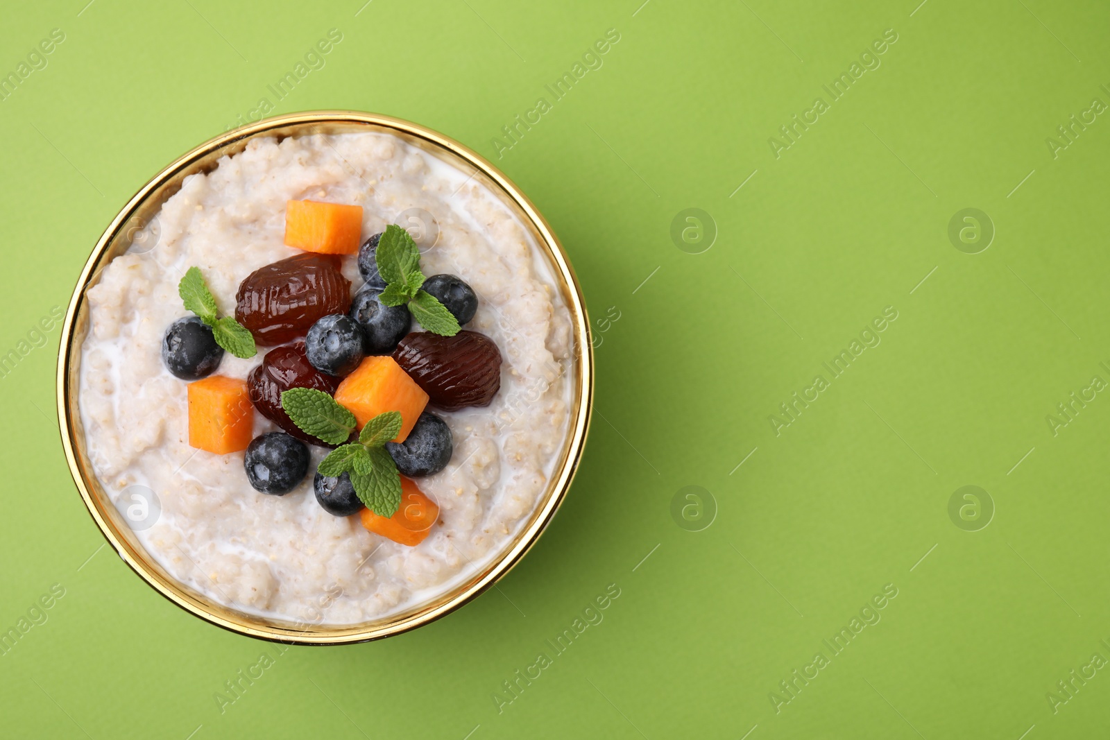 Photo of Delicious barley porridge with blueberries, pumpkin, dates and mint in bowl on green table, top view. Space for text