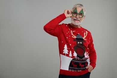 Senior man in Christmas sweater and funny glasses on grey background. Space for text