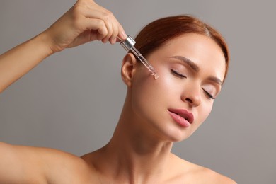 Beautiful young woman applying cosmetic serum onto her face on grey background