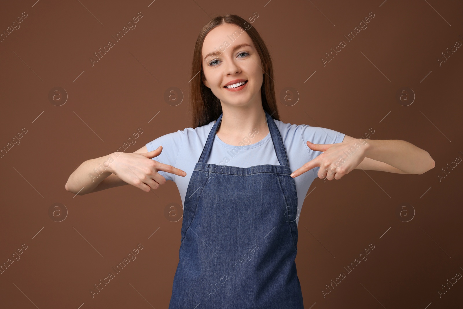 Photo of Beautiful young woman pointing at kitchen apron on brown background. Mockup for design