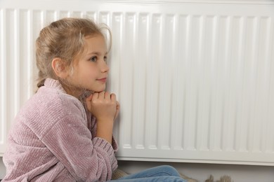 Little girl near heating radiator at home, space for text