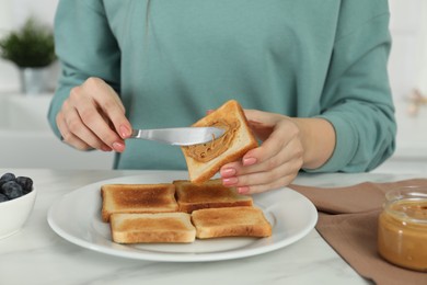 Woman spreading tasty nut butter onto toast at white table, closeup