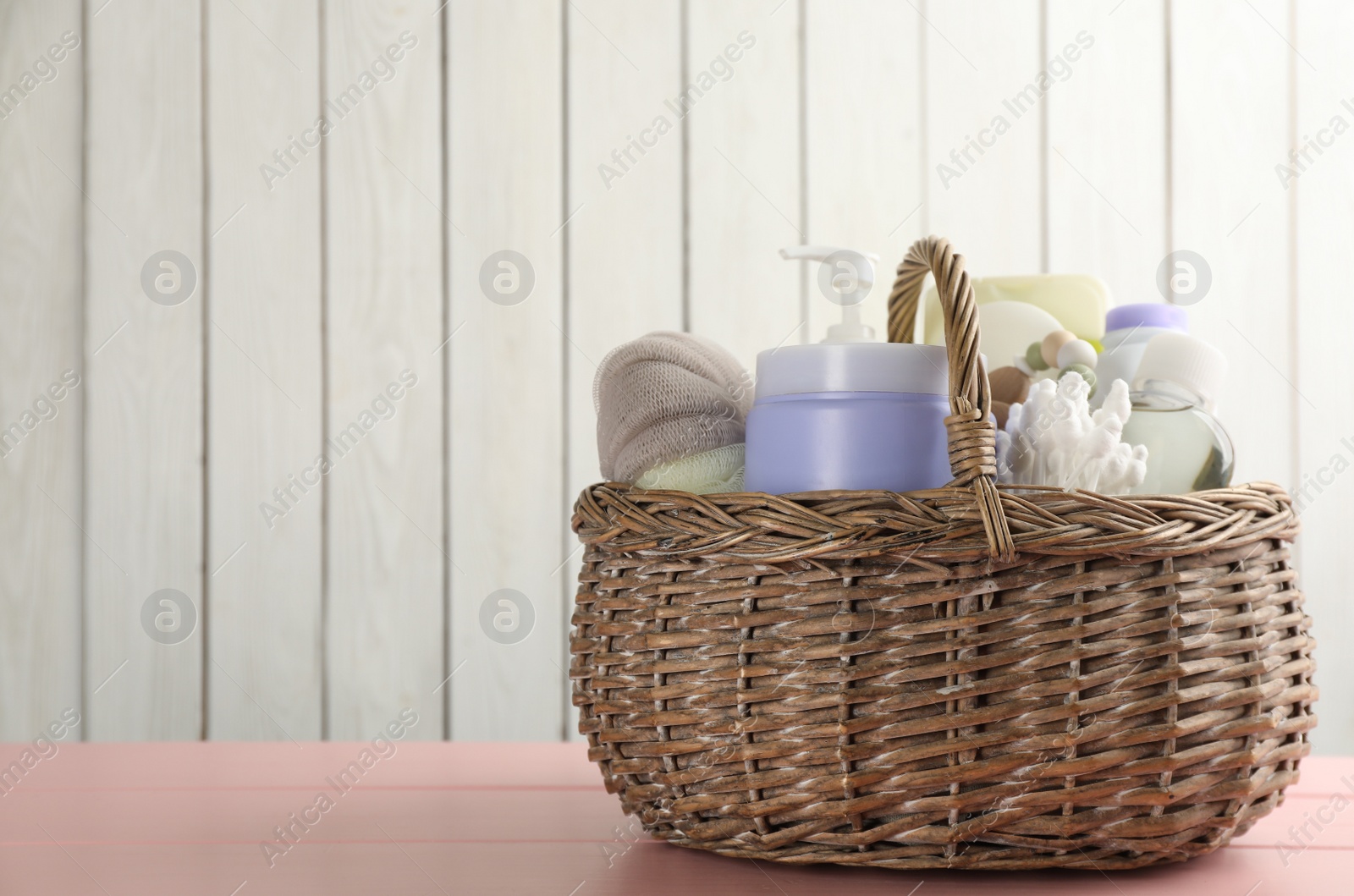 Photo of Wicker basket full of different baby cosmetic products and bathing accessories on pink wooden table. Space for text