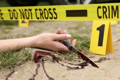 Crime scene with dead woman's body, bloody knife, marker and yellow tape outdoors, closeup