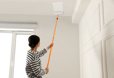 Young woman painting ceiling with white dye indoors, space for text