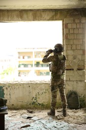Military mission. Soldier in uniform with binoculars inside abandoned building, back view