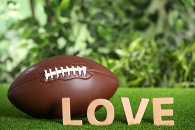 Photo of American football ball and word Love on green grass against blurred background. Space for text