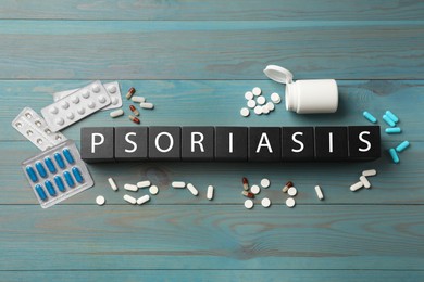 Word Psoriasis made of black cubes with letters and pills on blue light wooden table, flat lay