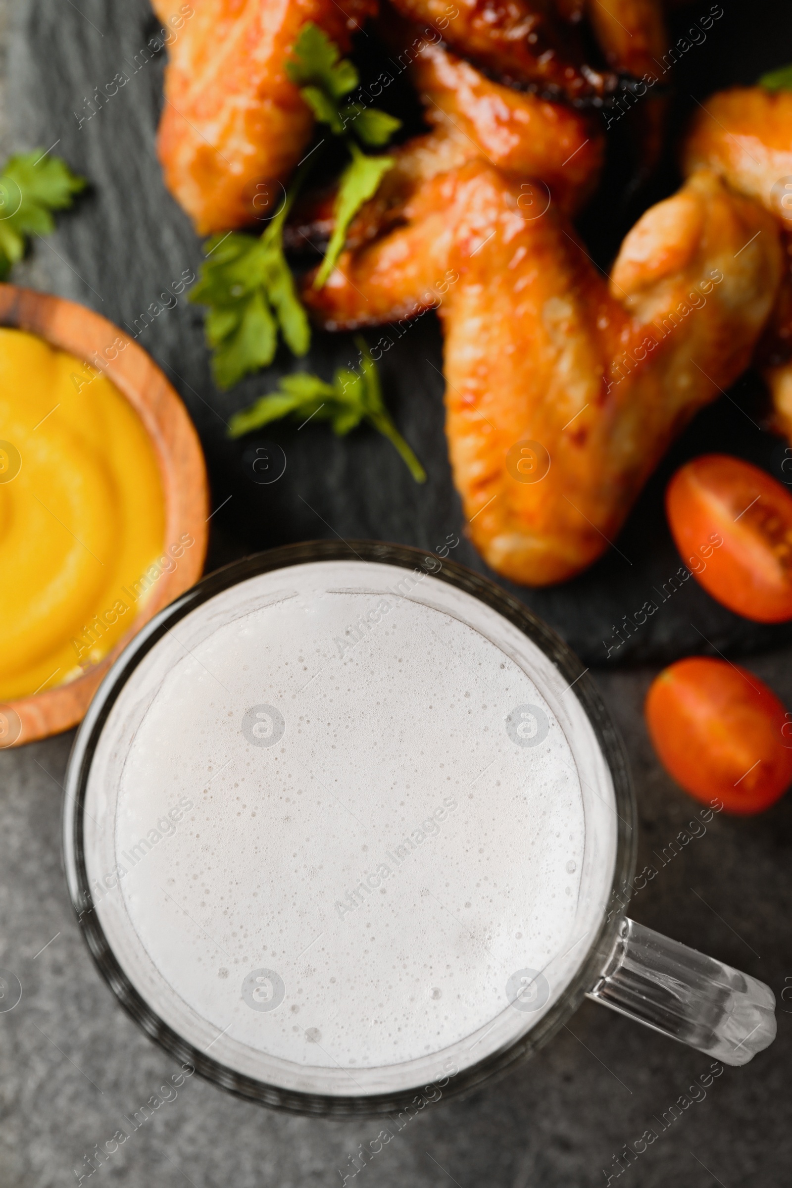 Photo of Mug with beer, delicious baked chicken wings and sauce on grey table, flat lay