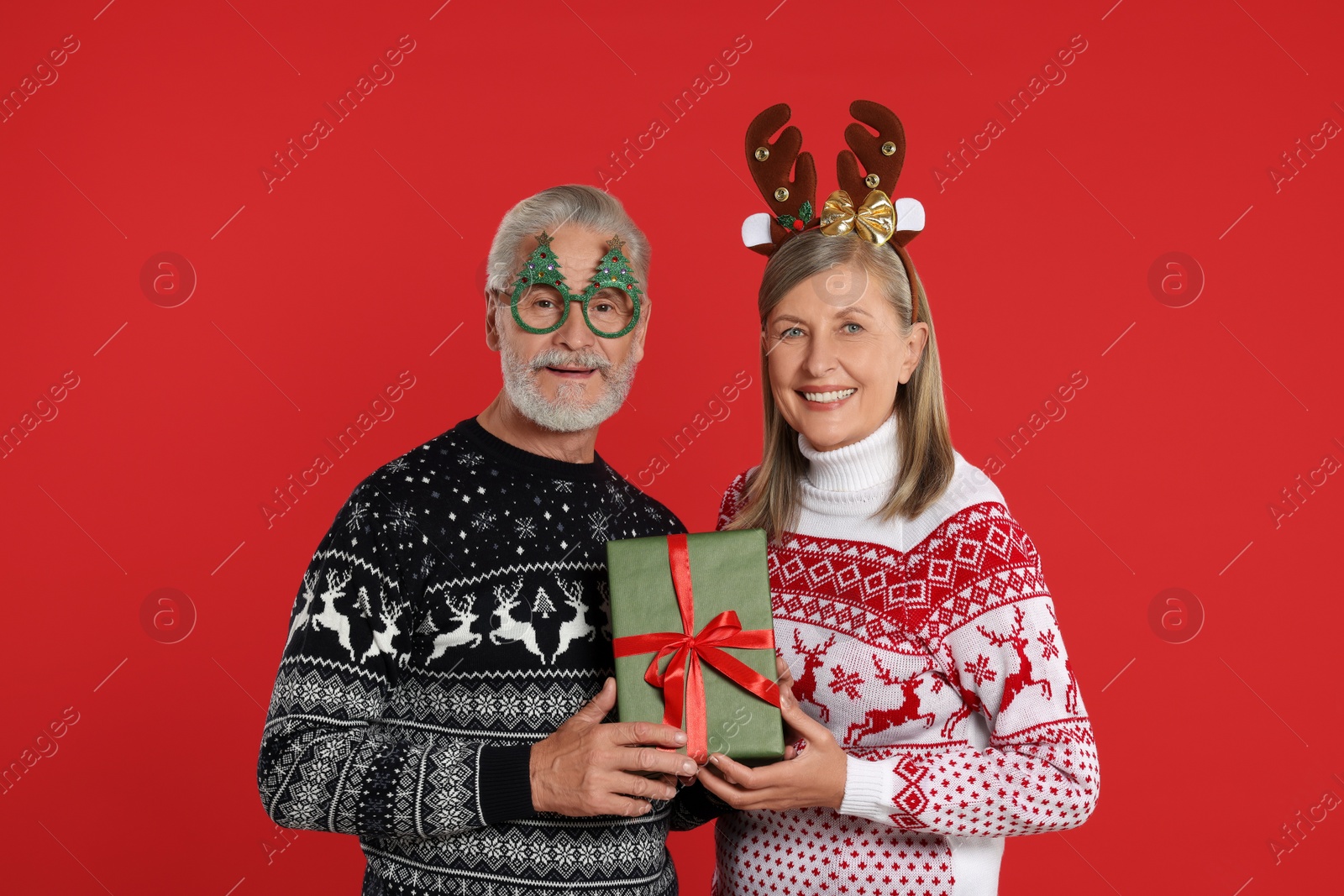 Photo of Senior couple in Christmas sweaters, reindeer headband and funny glasses holding gift on red background