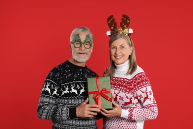 Photo of Senior couple in Christmas sweaters, reindeer headband and funny glasses holding gift on red background