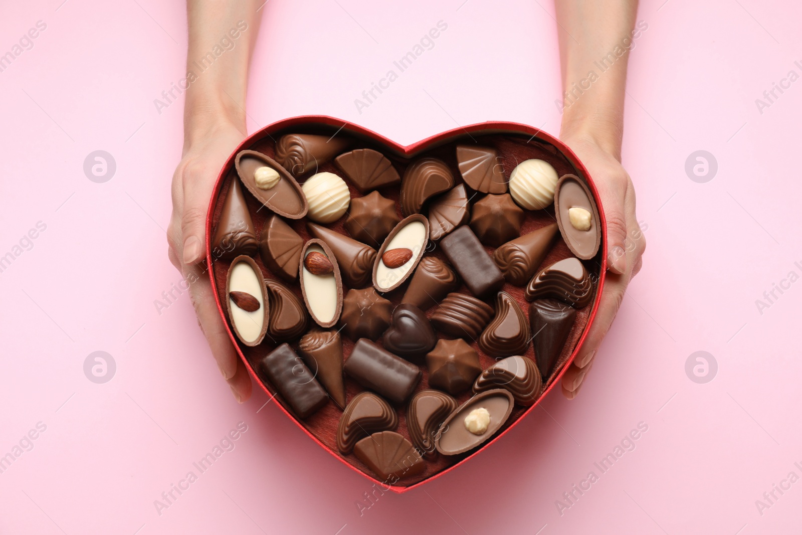 Photo of Woman holding heart shaped box with delicious chocolate candies on pink background, top view
