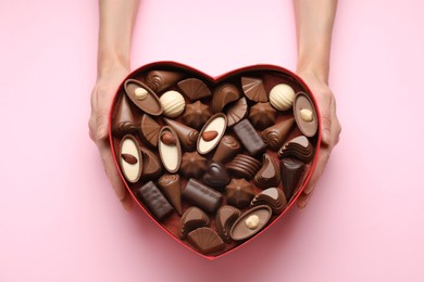 Woman holding heart shaped box with delicious chocolate candies on pink background, top view