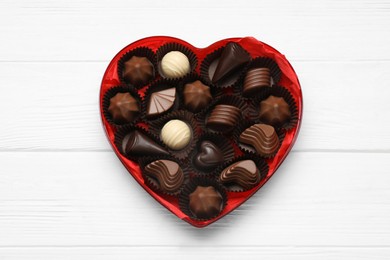 Photo of Heart shaped box with delicious chocolate candies on white wooden table, top view