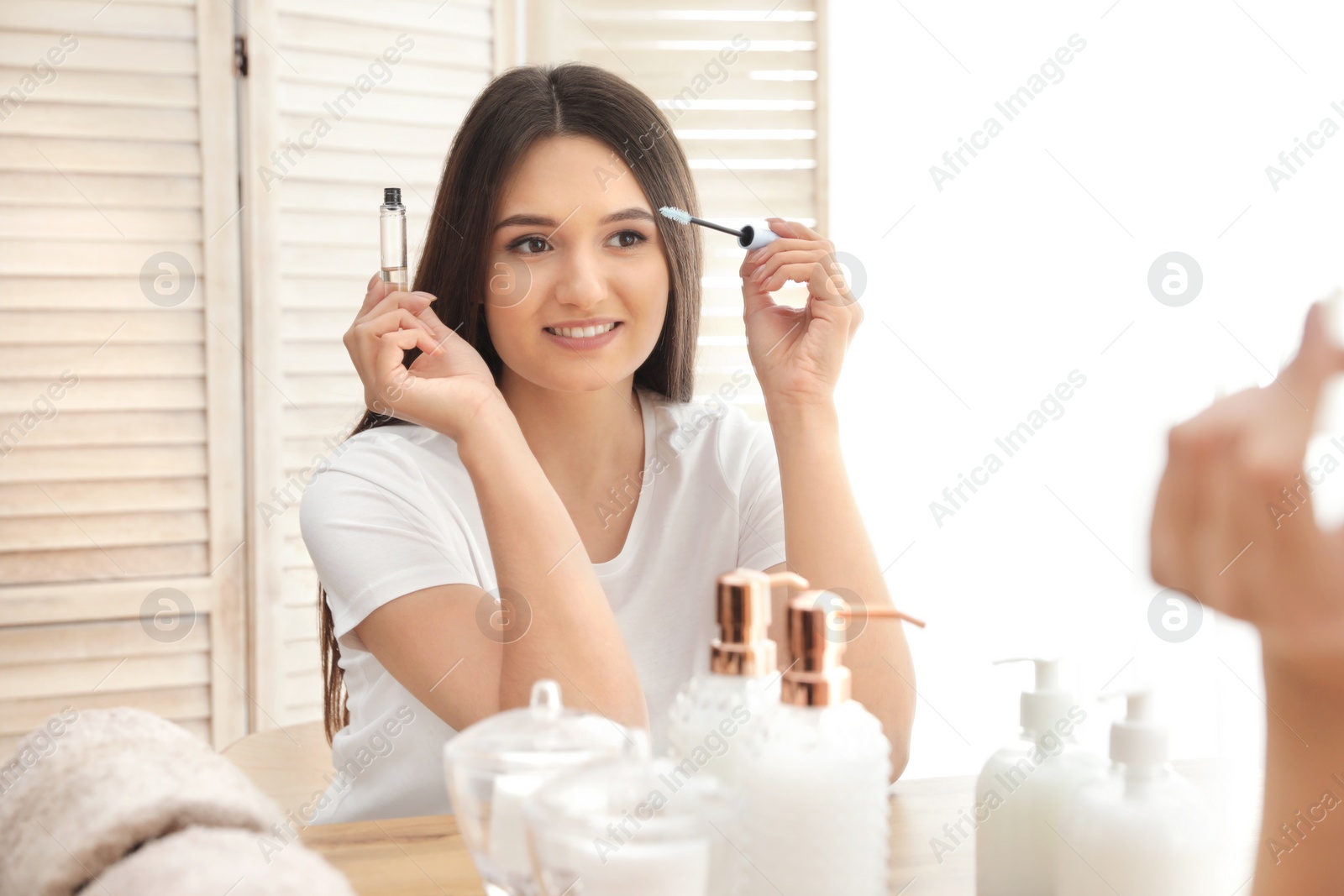 Photo of Beautiful woman applying oil onto her eyelashes near mirror indoors