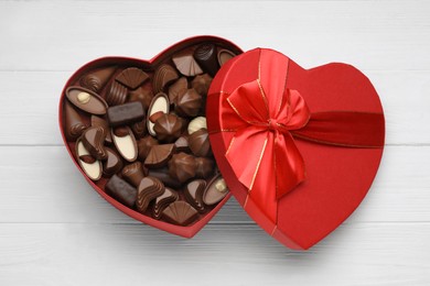 Photo of Heart shaped box with delicious chocolate candies on white wooden table, top view