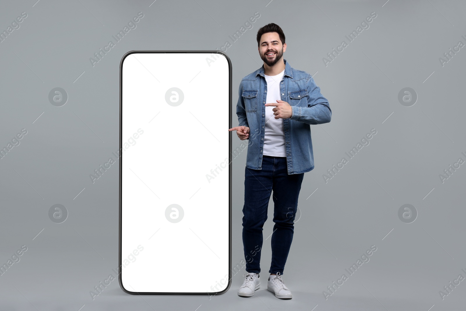 Image of Man pointing at huge mobile phone with empty screen on grey background. Mockup for design