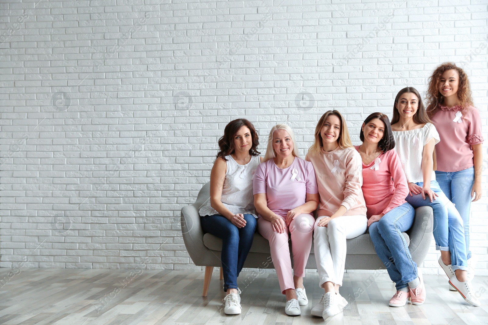Photo of Group of women with silk ribbons on sofa against brick wall, space for text. Breast cancer awareness concept