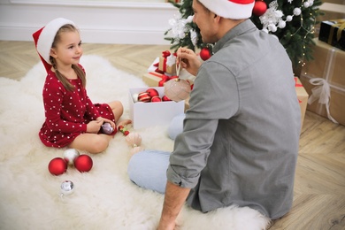 Father with his cute daughter in Santa hats decorating Christmas tree together at home