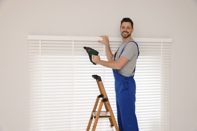 Photo of Worker in uniform installing window blind on stepladder indoors