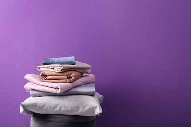 Photo of Stack of clean bed sheets and pillow on stool against purple background. Space for text