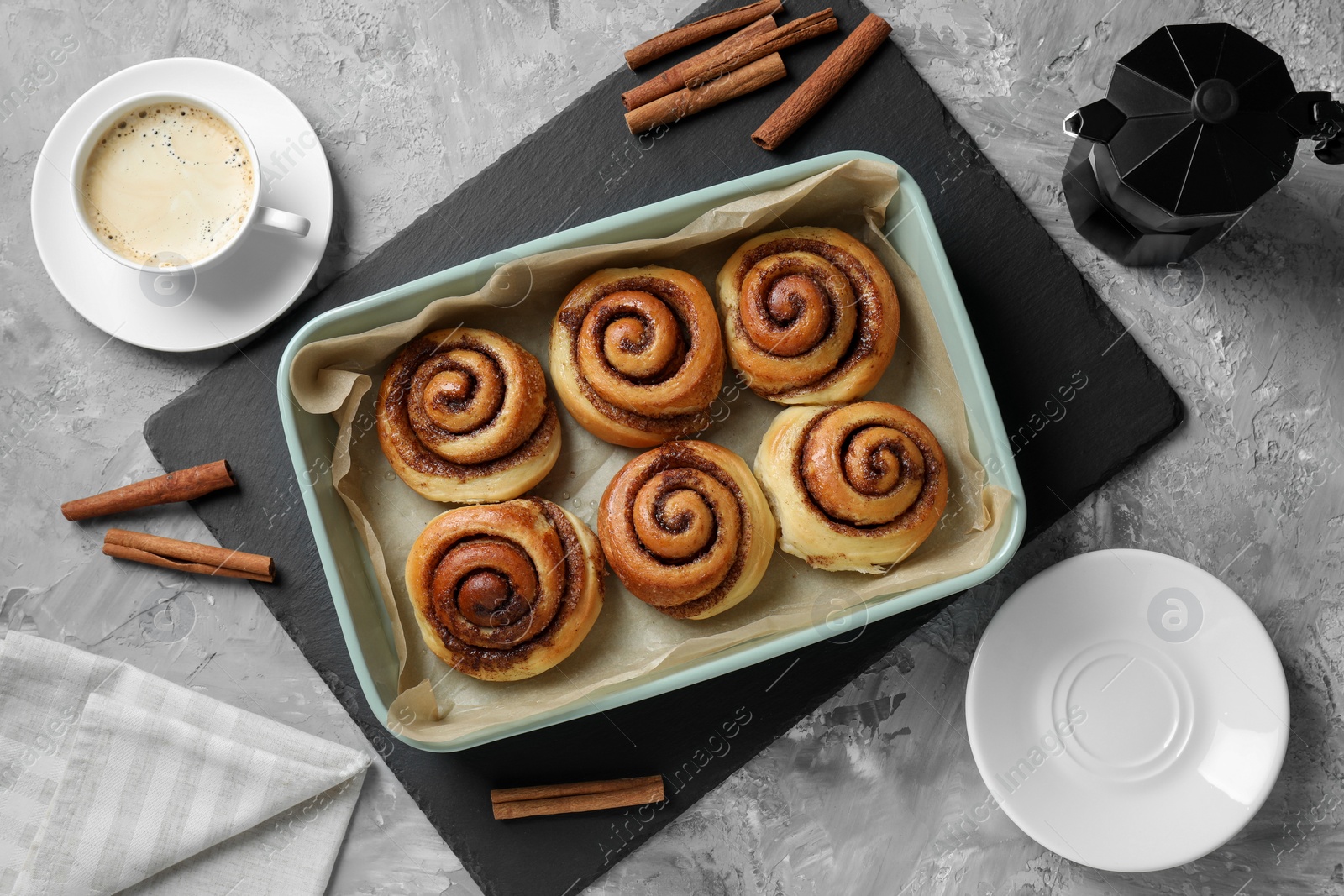 Photo of Baking dish with tasty cinnamon rolls, sticks and coffee on grey textured table, flat lay